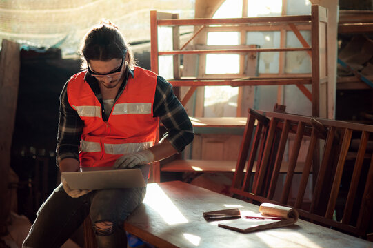 Young and handsome caucasian carpenter in the carpentry shop is working and checking customer's order in laptop to repair a furniture.small business and freelance craftsman.