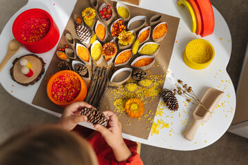 Child hands creating autumn tree with colored rice and natural materials. Toddler filled the leaves with yellow and orange rice. Montessori material. Sensory play and fall nature crafts