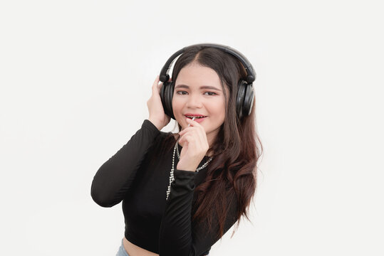 A cute Asian female poses for a studio picture with a white background while jamming to the music with her wireless headphones and eating lollipop.
