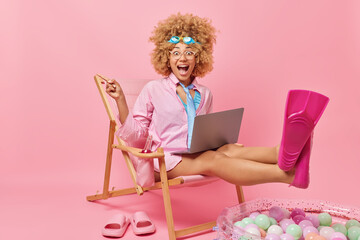 Horizontal shot of curly woman with amazed expression points index finger into distance works on laptop freelance wears goggles formal shirt and flippers sits on deck chair pretends being on beach © WHstudio Leushin N