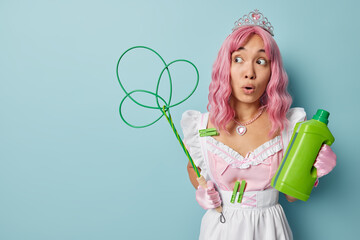 Impressed housewife with pink hair holds carpet beater and bottle of detergent reacts on something amazing wears crown and dress with stuck clothespins isolated over blue background empty space