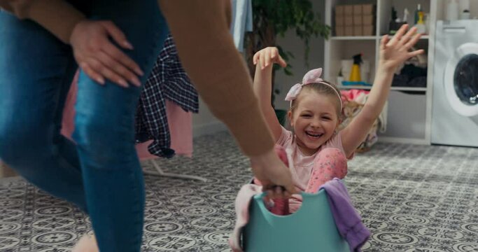 Sunny day. Mother and daughter were doing laundry. It's time for games and woman puts girl in washing bowl and rolls around the bathroom. Little girl in pink dress laughs loudly and waves hands.