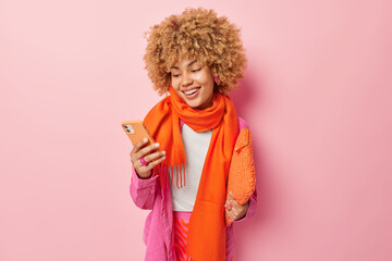 Positive curly haired woman smiles broadly sends text messages via smartphone carries small bag under arm feels glad wears fashionable shirt and orange scarf around neck isolated on pink wall