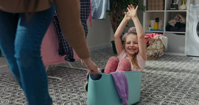 Older sister is playing with younger sister. She puts girl in the wash basin and rolls around the bathroom. Six-year-old girl laughs loudly and waves hands. Washed clothes hang behind.