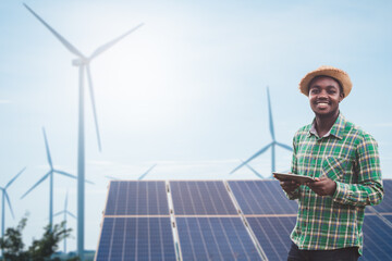 African farmer standing and holding digital tablet on corn farm with solar cell and wind turbine in...