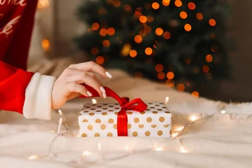 The concept of Christmas. Christmas gift box on the background of a Christmas tree decorated with garlands. A woman's hand with a manicure opens a gift in anticipation of the holiday. No people 