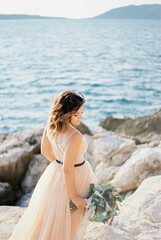 Fototapeta na wymiar Bride with a bouquet stands sideways with her head bowed on a rocky seashore