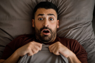 A man in a burgundy T-shirt is scared in bed looking at the camera