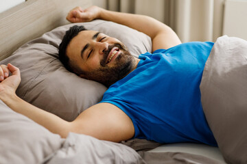 Happy middle eastern man stretching in bed after a good sleep, relaxing in the bedroom, copy space