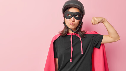 Horizontal shot of attentive superwoman raises arms shows strong biceps ready to save world dressed in superhero costume has thoughtful expression isolated over pink background blank space on left