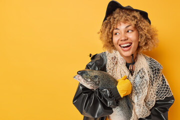 Cheerful angler poses with big stout looks away with happy expression smiles broadly carries fishing net dressed in comfortable clothes isolated over yellow background blank space for your text