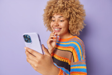 Pretty cheerful woman with curly hair holds smartphone in front of face has pleasant online talk...