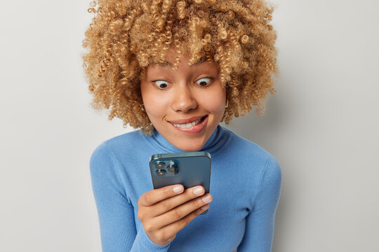 Image of surprised curly haired woman looks amazed at smartphone screen bites lips cannot believe own eyes finds out shocking news wears casual blue turtleneck poses indoors against grey wall