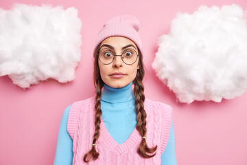 Beautiful amazed teenage girl with two pigtails stares through transparent eyeglasses cannot believe own eyes reacts to shocking news wears hat turtleneck and vest poses indoor white clouds around