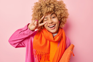 Photo of cheerful woman makes v sign gesture over eye smiles toothily expresses positive emotions wears fashionable clothes carries small bag isolated on pink background. Body language concept