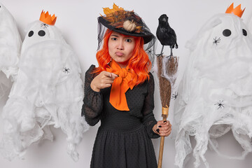 Happy Halloween concept. Serious displeased witch poses with broom and crow poits index finger at you reproaches about something prepares for holiday carnival at end of October. Autumn and celebration