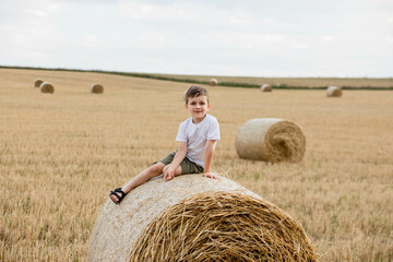 Naklejka na ściany i meble A cute little boy in a white t-shirt and khaki shorts is sitining on round bales of hay. Photo session in the field