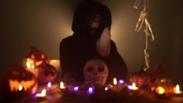 Blur portrait of a praying wizard among Halloween decorations on black background. A young caucasian man in a dark mantle is conjuring holding hands together surrounded by jack o'lanterns and a skull.