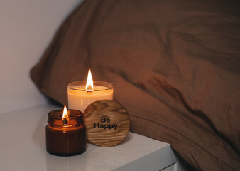 A burning candle in a jar near the bed with a wooden inscription "Be happy". The concept is cozy.