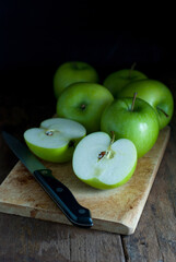 Green apples, whole, and halves with a kitchen knife put on a wooden cutting board on a rustic wooden floor.