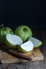 Slice and whole guava fruit with a kitchen knife and wooden cutting board on a rustic wooden background.