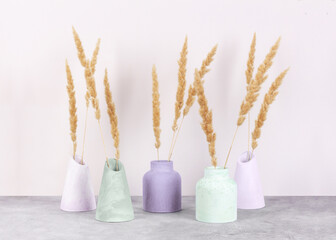 Five multicolored plaster vases with dried flowers on a gray concrete table against the pale pink wall, front view. Home decor.