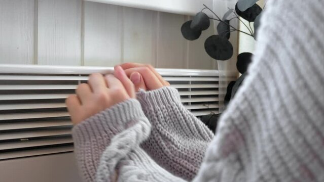 Young frozen woman wearing a sweater shaking and freezing for winter cold with shock expression on face. Girl is warming up hands with breath over electric heater. Discomfort spending time at home