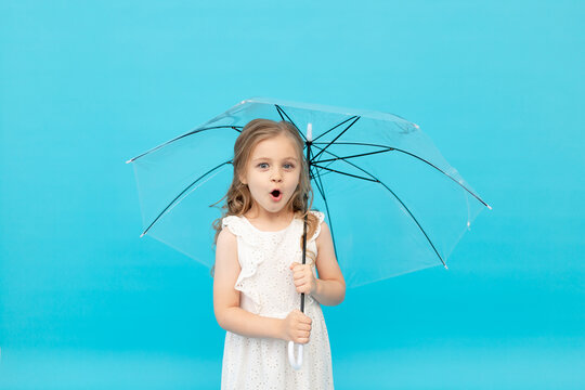 surprised cute little girl in a cotton white dress holding a transparent umbrella on a blue background in the studio, space for text