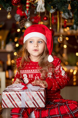 Fototapeta na wymiar a little cute girl child in a red sweater and a Santa Claus hat in a dark kitchen with a Christmas tree rejoices with gifts and waits for the new year or Christmas