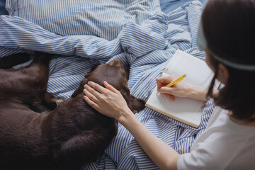 top view, woman in bed writes a diary or notes about her dreams, plans for the day. A European female writer writes a letter and hugs a Labrador retriever dog. A diary of emotions and notes about your