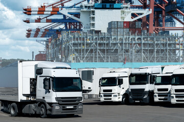 Obraz premium Trucks in the international seaport against on a background of a ship loaded with containers. International trade and logistics concept