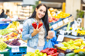 Beautiful smiling woman selects two red pepper from a wide assortment of fresh fruit and veg in the market