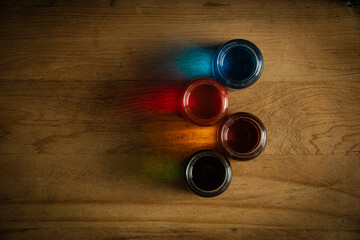 Four glass jars on an old wooden table that contain water with paint and the light reflects the color on the wood