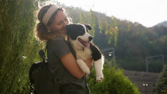 a girl with a border collie puppy in her arms kisses him and hugs him by a tree in the sunlight against the background of mountains