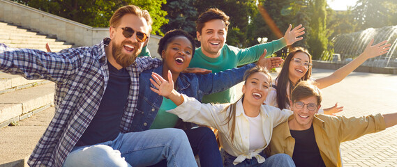 Happy cheerful young positive multiracial friends posing for group photo in a summer park sitting on steps and spreading hands wide in sides smiling looking at camera. Friendship, having fun concept.