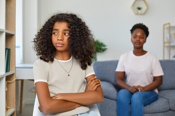 Attractive teenage girl is looking sadly away. Girl sits with her arms crossed in closed pose. Defocused Mom sitting behind and looks at her daughter. African american family at home. - obrazy, fototapety, plakaty