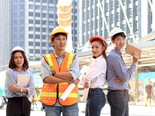 Portrait of four smart young Asian man and woman engineer group, staff worker team wear safety vest and helmet, confidential standing at outside office in city with skyscraper building as background.