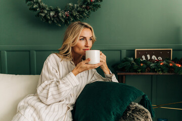 A woman in pajamas drinks a hot drink while sitting in a living room in a Christmas decor.