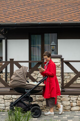 Young mother in a red coat walks with a stroller and reads news on her phone.