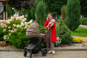 Stylish mother in a red coat with a baby in her arms walks with a stroller and talks on the phone.