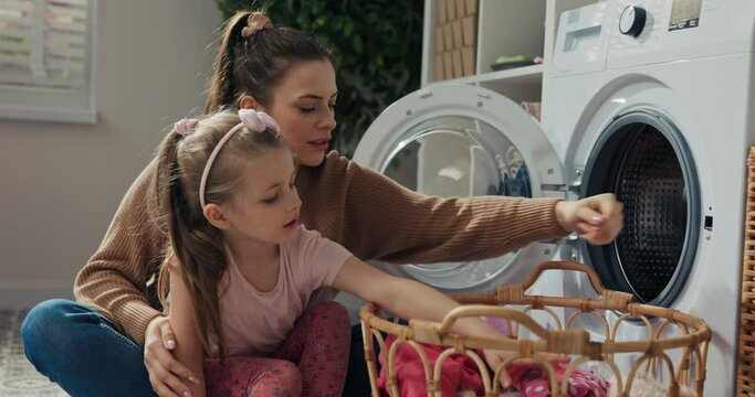 There are dirty clothes in laundry basket. Smiling girl and mother throw clothes into washing machine. They take clothes one by one and chat. Girl is wearing pink clothes, and mother brown sweater.