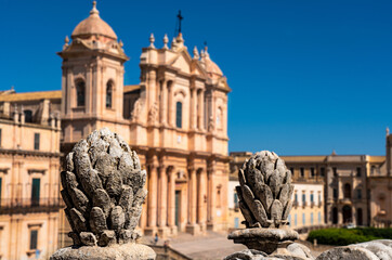 Fototapeta na wymiar In the foreground baroque decorations in stone in the shape of a pine cone that frame the facade of the cathedral of Noto placed in the background out of focus