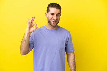 Handsome blonde man over isolated yellow background showing ok sign with fingers