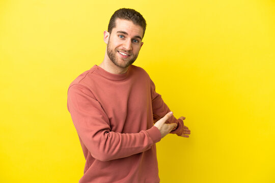 Handsome blonde man over isolated yellow background extending hands to the side for inviting to come