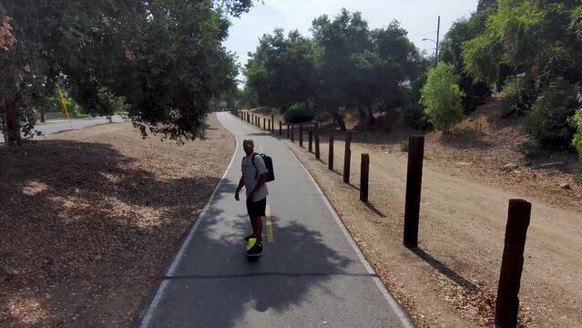 aerial footage of an African American man riding a Onewheel electric skateboard on a bike trail surrounded by lush green trees and grass and dry brush with blue sky at Duarte California USA