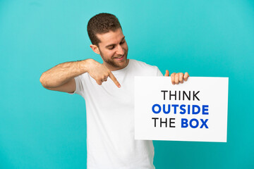 Handsome blonde man over isolated blue background holding a placard with text Think Outside The Box and  pointing it