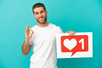 Handsome blonde man over isolated blue background holding a placard with Like icon and celebrating a victory