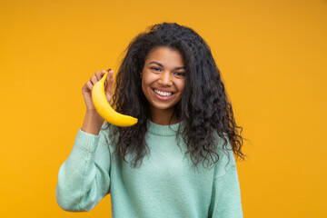 Studio close up portrait of cute smiling african american girl posing with fresh ripe banana in hand, isolated over bright colored yellow backgorund