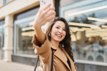 Glamour attractive asian kazakh female wearing beige coat, taking selfie, checking haircut, look at smartphone camera, walking on street
