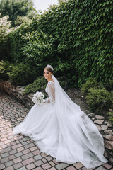 A beautiful bride in a long white dress with a bouquet in her hands, a diadem on her head, sits in the park, against the background of a wall with green ivy. Wedding photography, portrait.